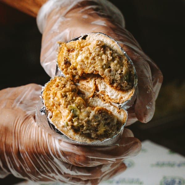 Gloved hands hold a foil-wrapped halal sandwich, cut in half to show the meaty, cheesy filling.
