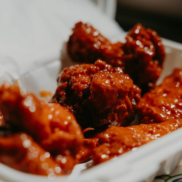 Close-up of several saucy, red halal chicken wings in a white takeout container.