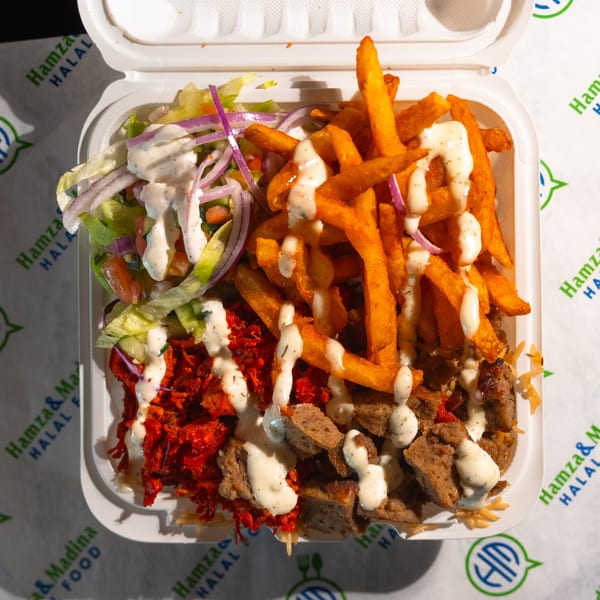 Halal food platter with chicken, lamb, fries, and salad, drizzled with white sauce in a takeout container.