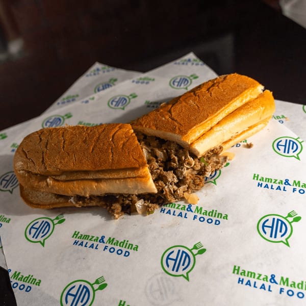 A Hamza & Madina Halal Food cheesesteak sandwich, cut in half, on branded wax paper.