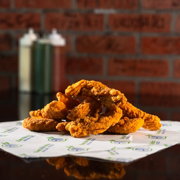 Crispy golden halal chicken tenders piled on branded paper with sauces and a brick wall in the background.