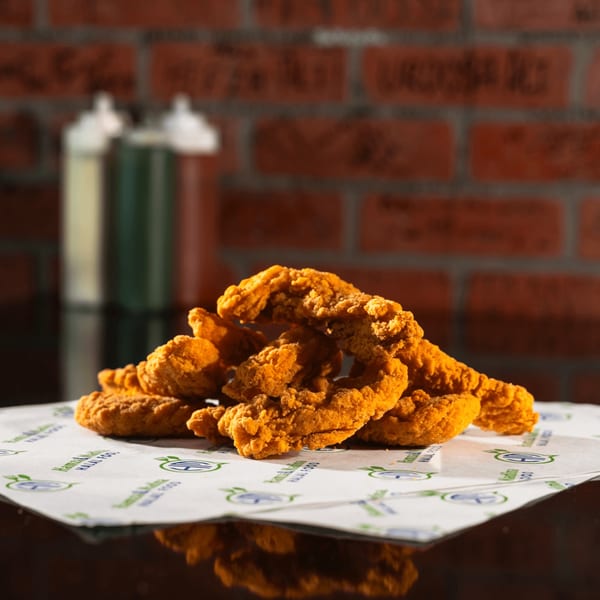 Crispy golden-brown halal chicken tenders piled on branded paper, with sauce bottles and a brick wall in the background.