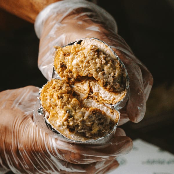 Gloved hands hold a foil-wrapped, sliced halal sandwich filled with meat and melted cheese.